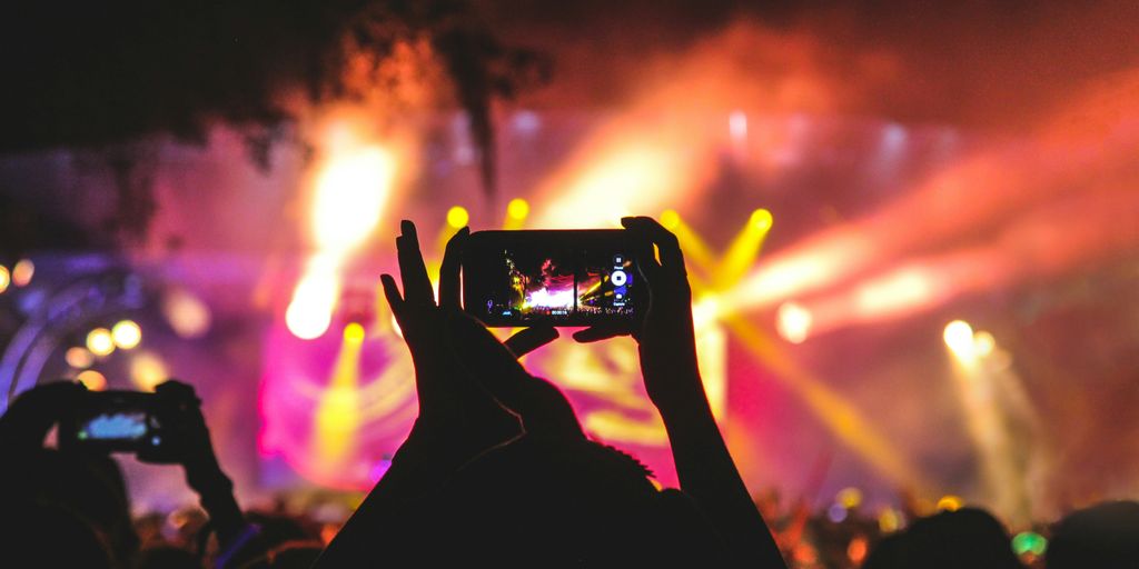 person holding smartphone taking video of a concert near stage with lights during nighttime